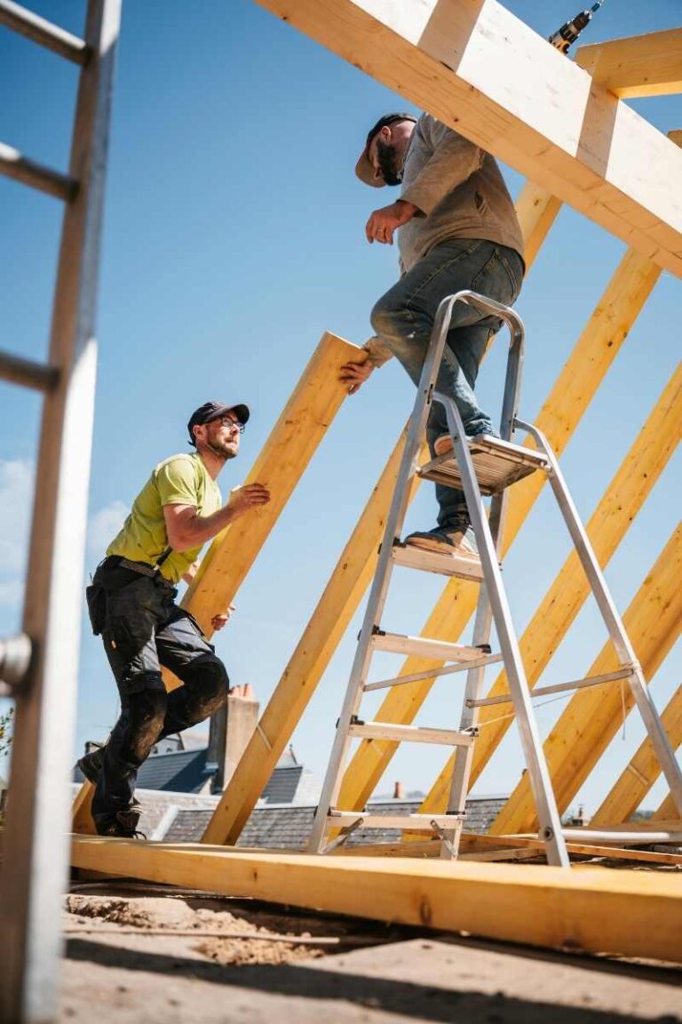 Two construction workers assembling a wooden roof frame under a clear blue sky - ccinsfl.com