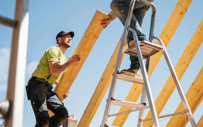 Two construction workers assembling a wooden roof frame under a clear blue sky - ccinsfl.com